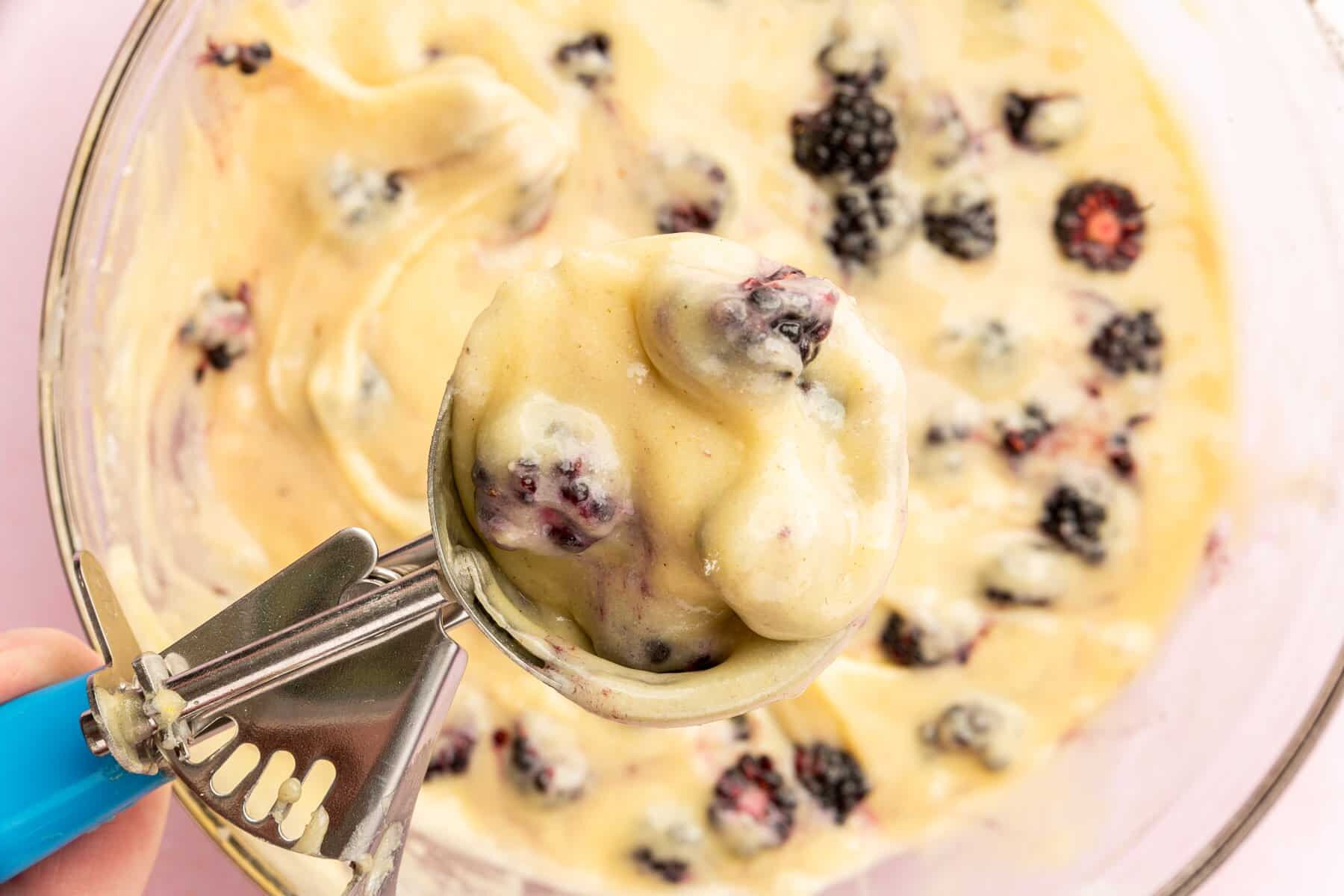 A closeup of a ice cream scoop filled with gluten-free blackberry muffin batter over a whole of more batter.