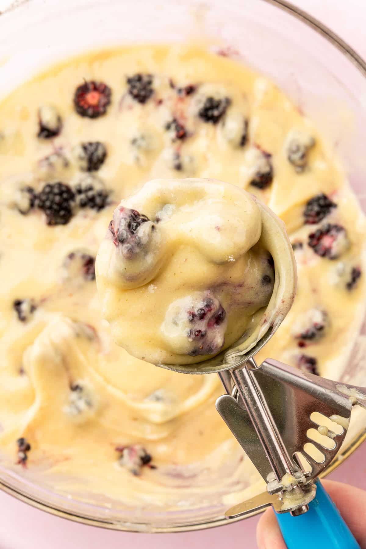A closeup of a ice cream scoop filled with gluten-free blackberry muffin batter over a whole of more batter.