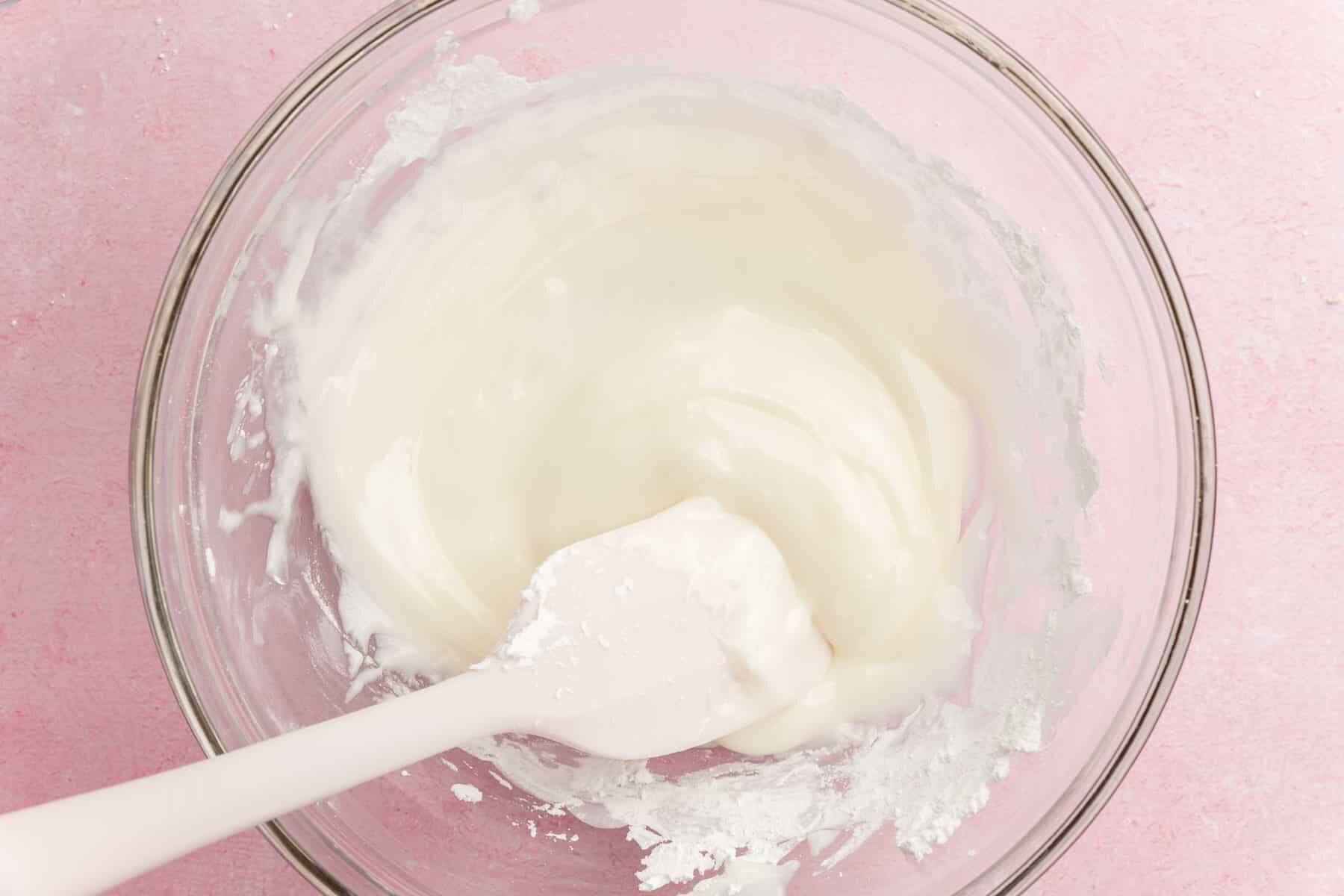 A glass bowl with white icing being mixed by a white spatula, set on a light pink surface.
