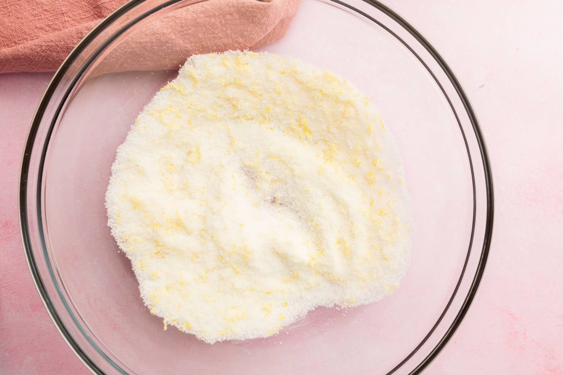 A glass mixing bowl with lemon zest and granulated sugar mixed together.