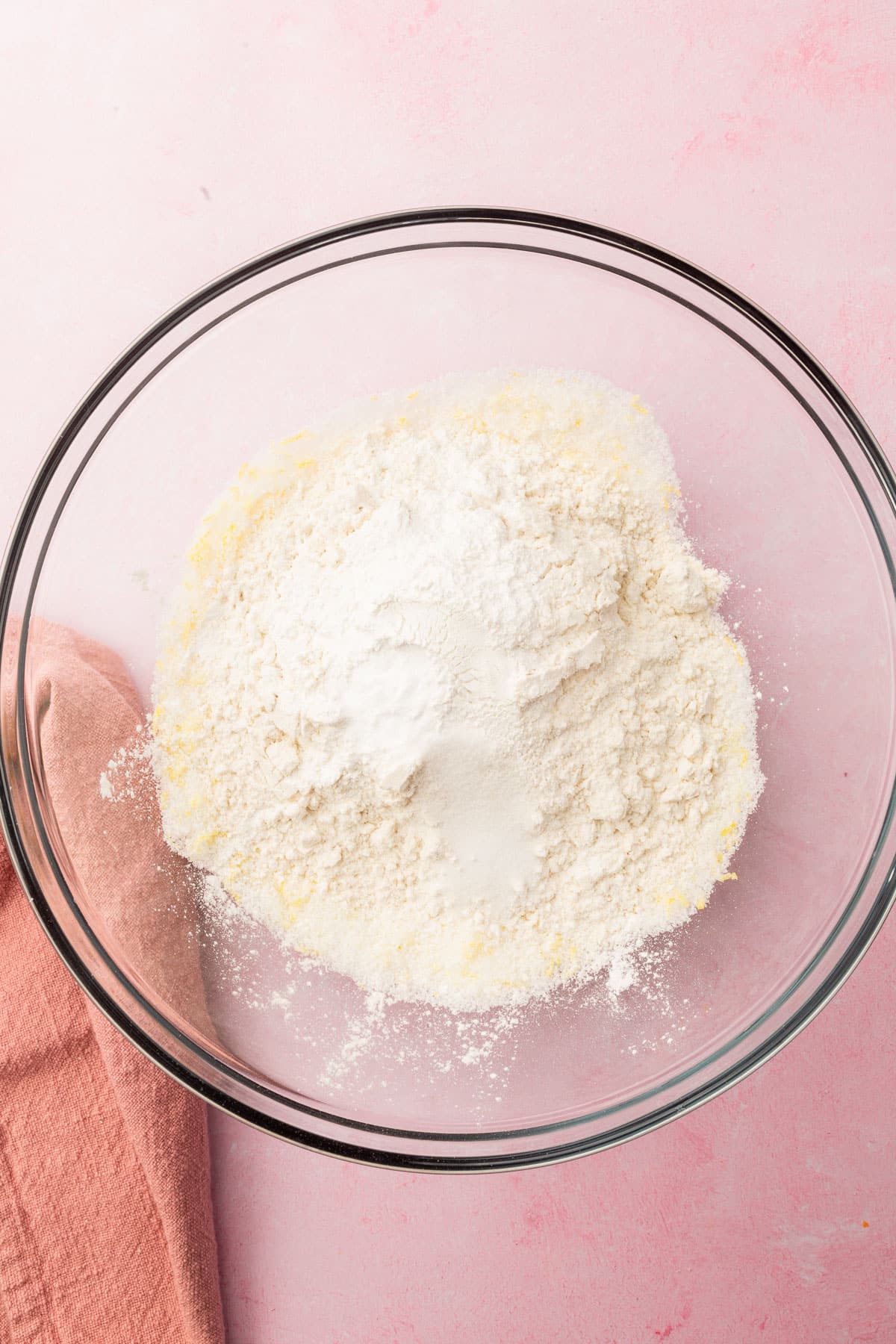 A glass mixing bowl with granulated sugar, lemon zest, gluten-free flour, baking powder, baking soda, and salt in it before mixing together.