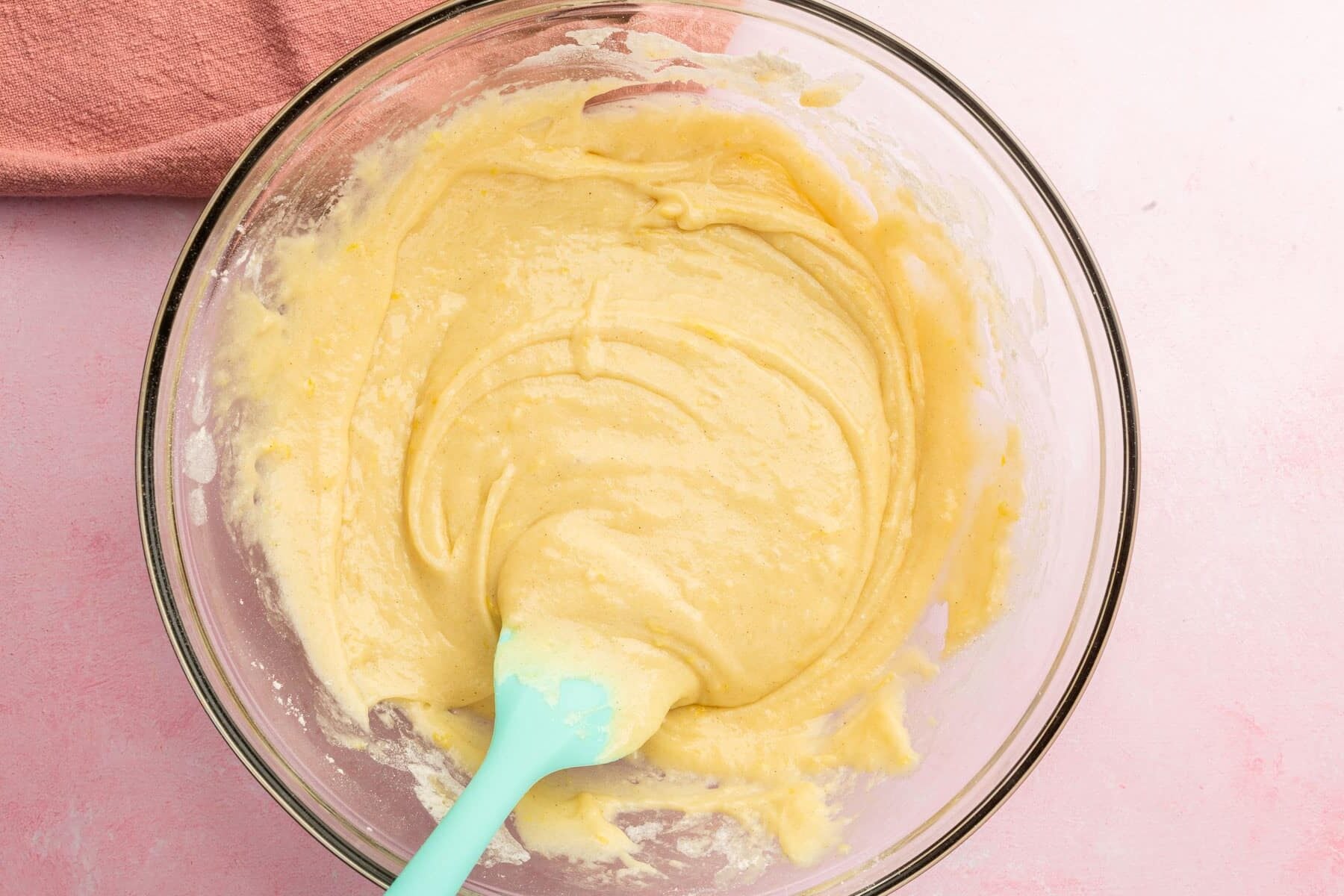 A glass mixing bowl with gluten-free muffin batter being stirred by a blue spatula.