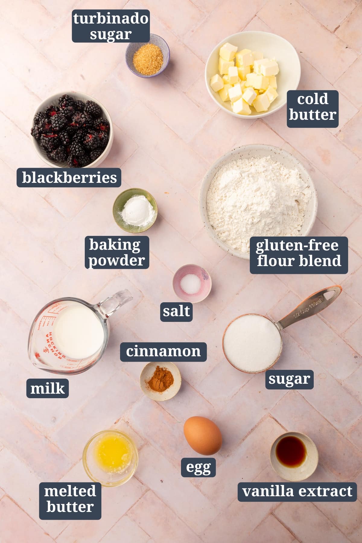Ingredients in small bowls to make gluten free blackberry scones including turbinado sugar, cold butter, blackberries, baking powder, gluten-free flour blend, salt, milk, cinnamon, sugar, egg, melted butter, and vanilla extract with text overlays over each ingreidnet.