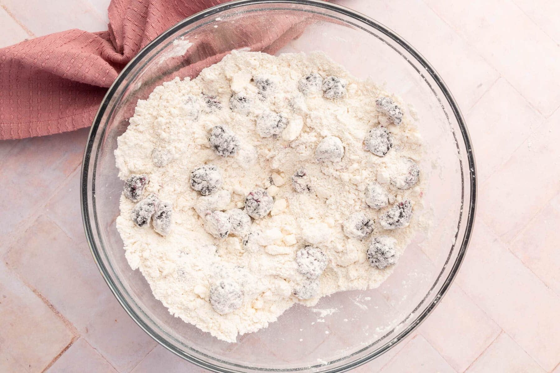 A glass mixing bowl with flour, little chunks of butter and blackberries that have all been mixed together.