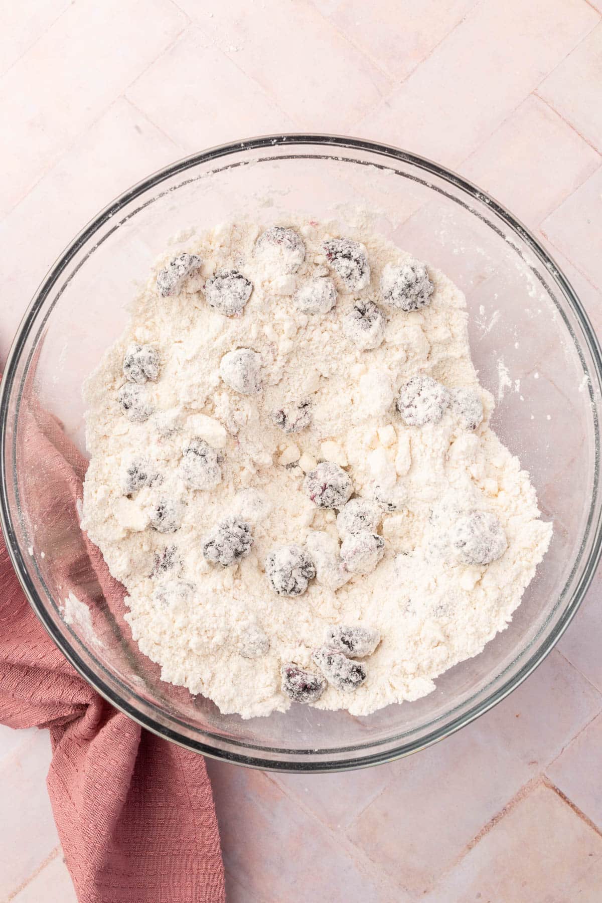 A glass mixing bowl with flour, little chunks of butter and blackberries that have all been mixed together.