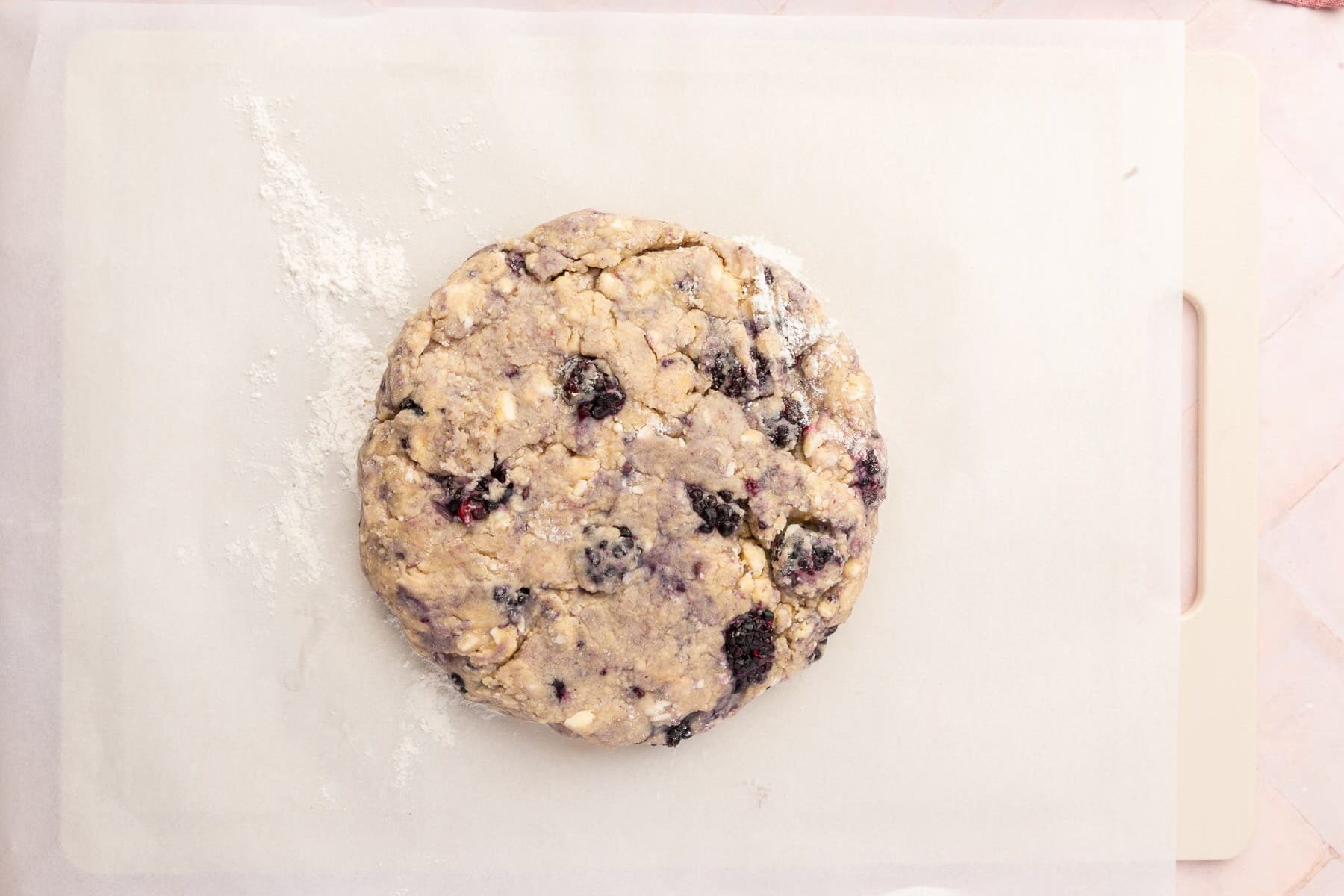 A round disk of gluten free blackberry scone dough on a cutting board lined with parchment paper.