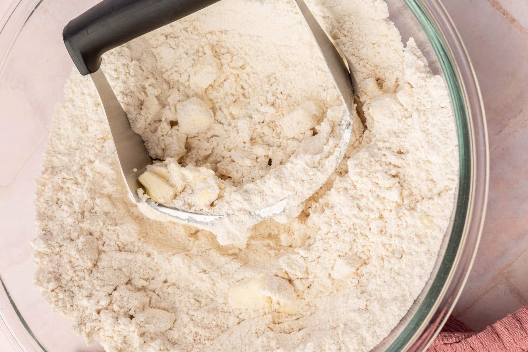 A glass mixing bowl with a pastry cutter that is cutting butter cubes into a gluten-free flour mixture.