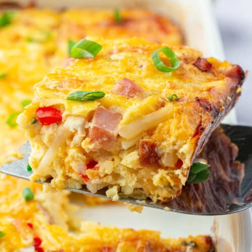 A spatula lifting a slice of ham egg and cheese casserole from the baking dish.