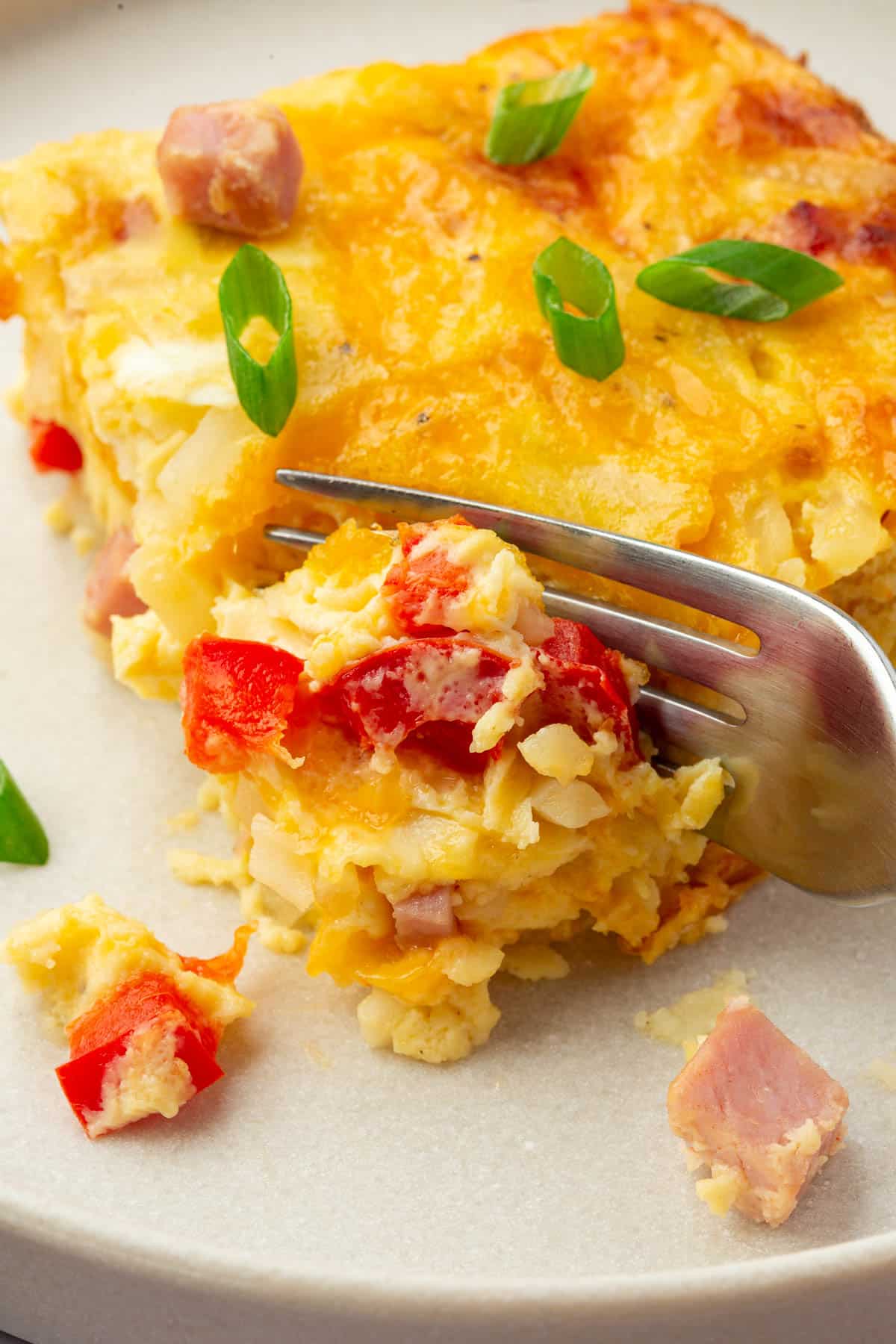 A fork cutting into a slice of gf breakfast casserole with ham, red bell pepper, and green onions.