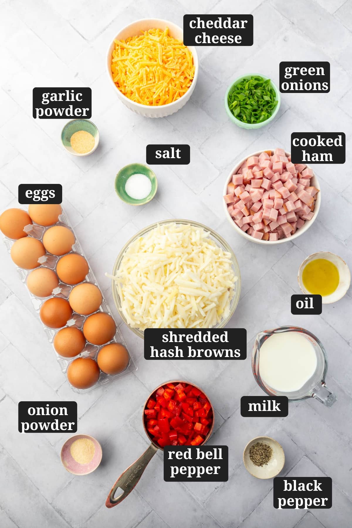 An overhead view of ingredients to make gluten free breakfast casserole including cheddar green, green onions, garlic powder, salt, cooked ham, eggs, shredded hash browns, oil, milk, red bell pepper, black pepper and onion powder with text overlays over each ingredient.