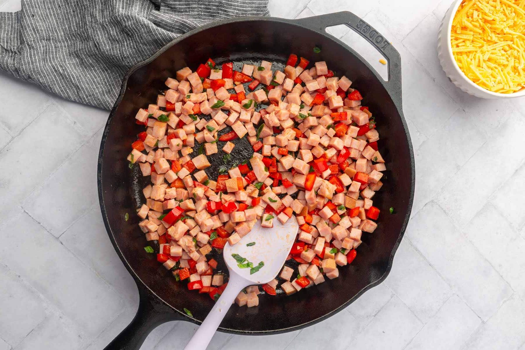 A cast iron skillet with sautéed cubed ham and red bell pepper with a spatula in it.