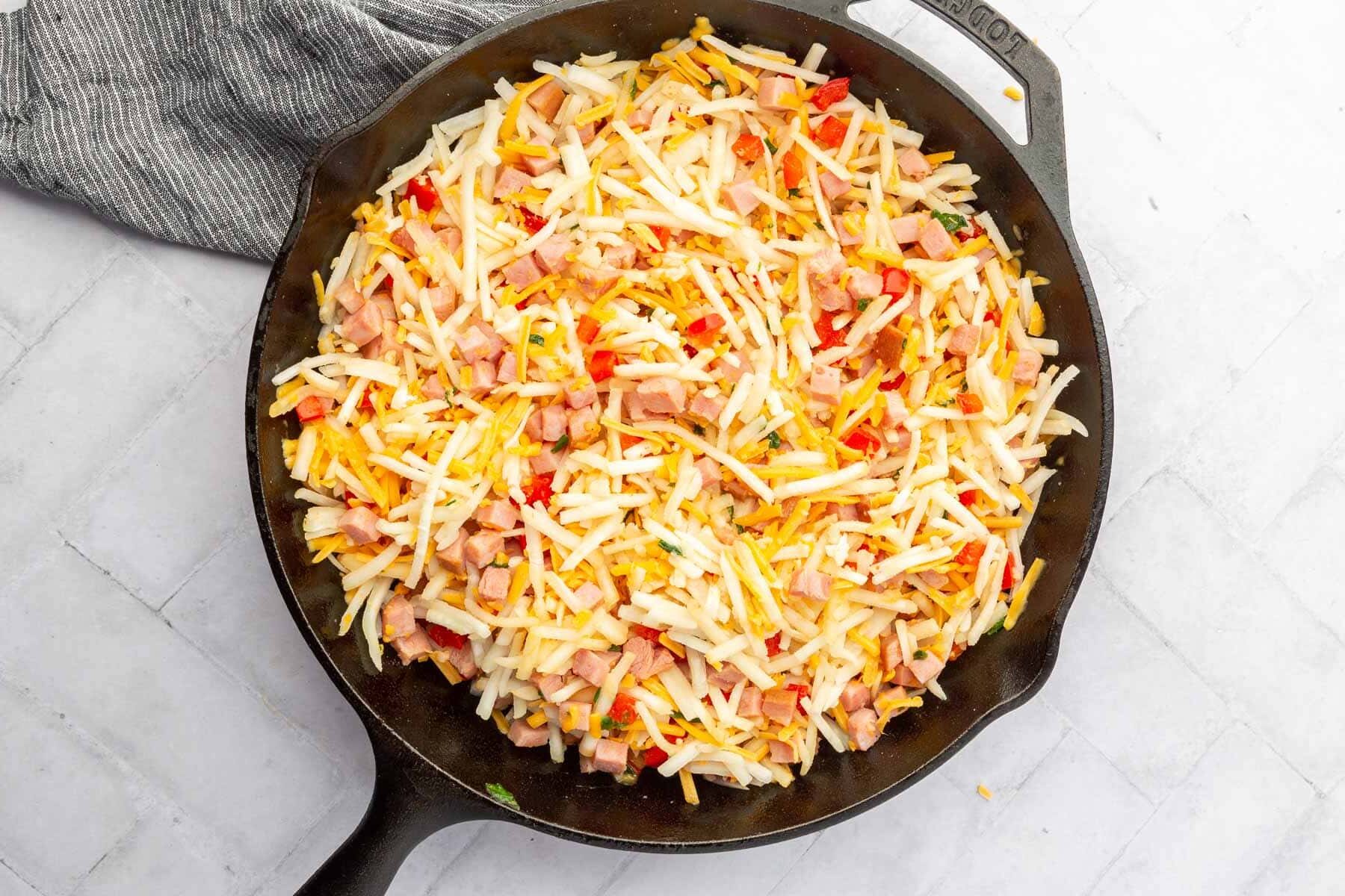 A cast iron skillet with hash browns, diced red bell pepper, shredded cheese, and cubed ham all mixed together.
