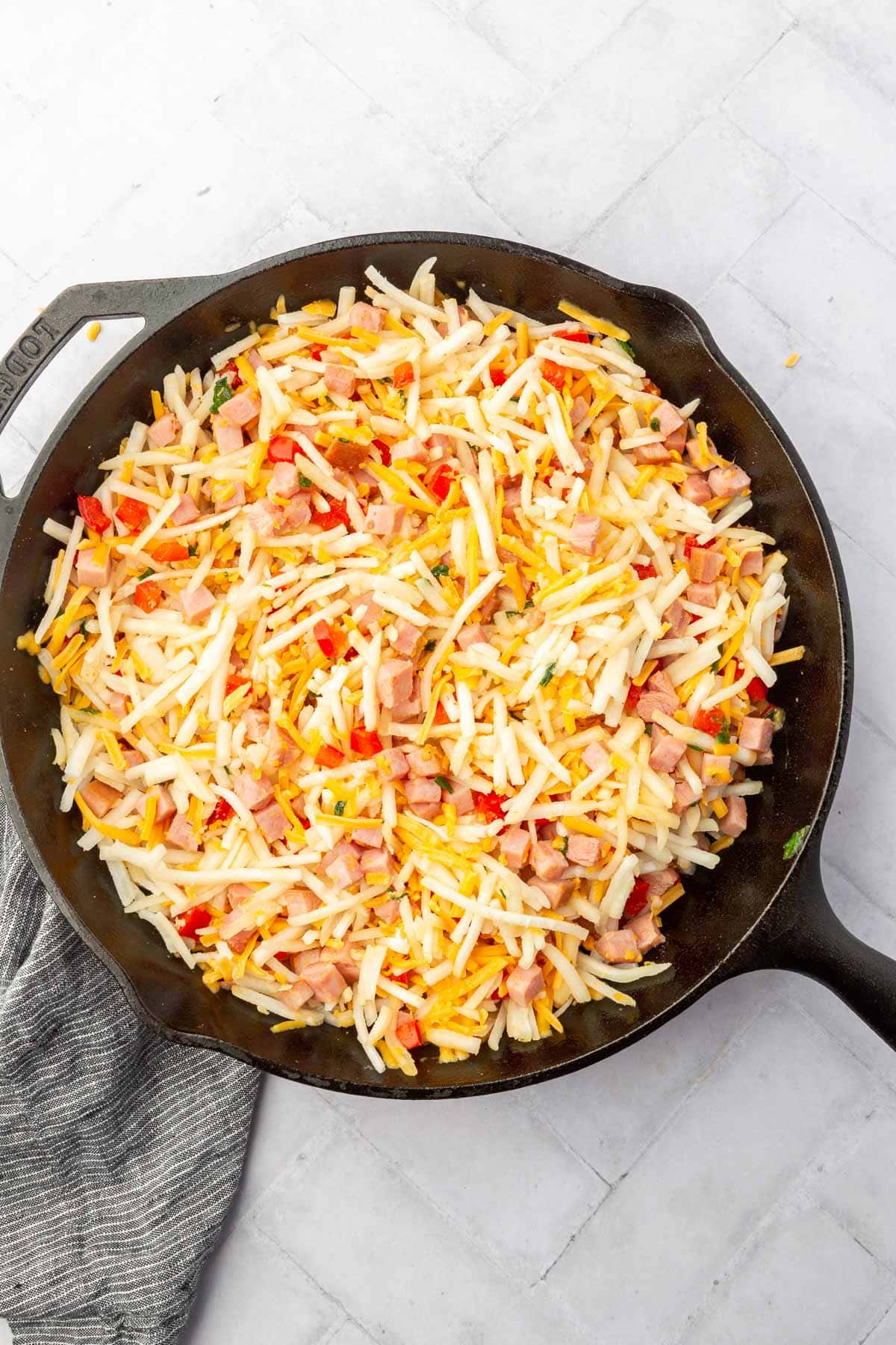 A cast iron skillet with hash browns, diced red bell pepper, shredded cheese, and cubed ham all mixed together.