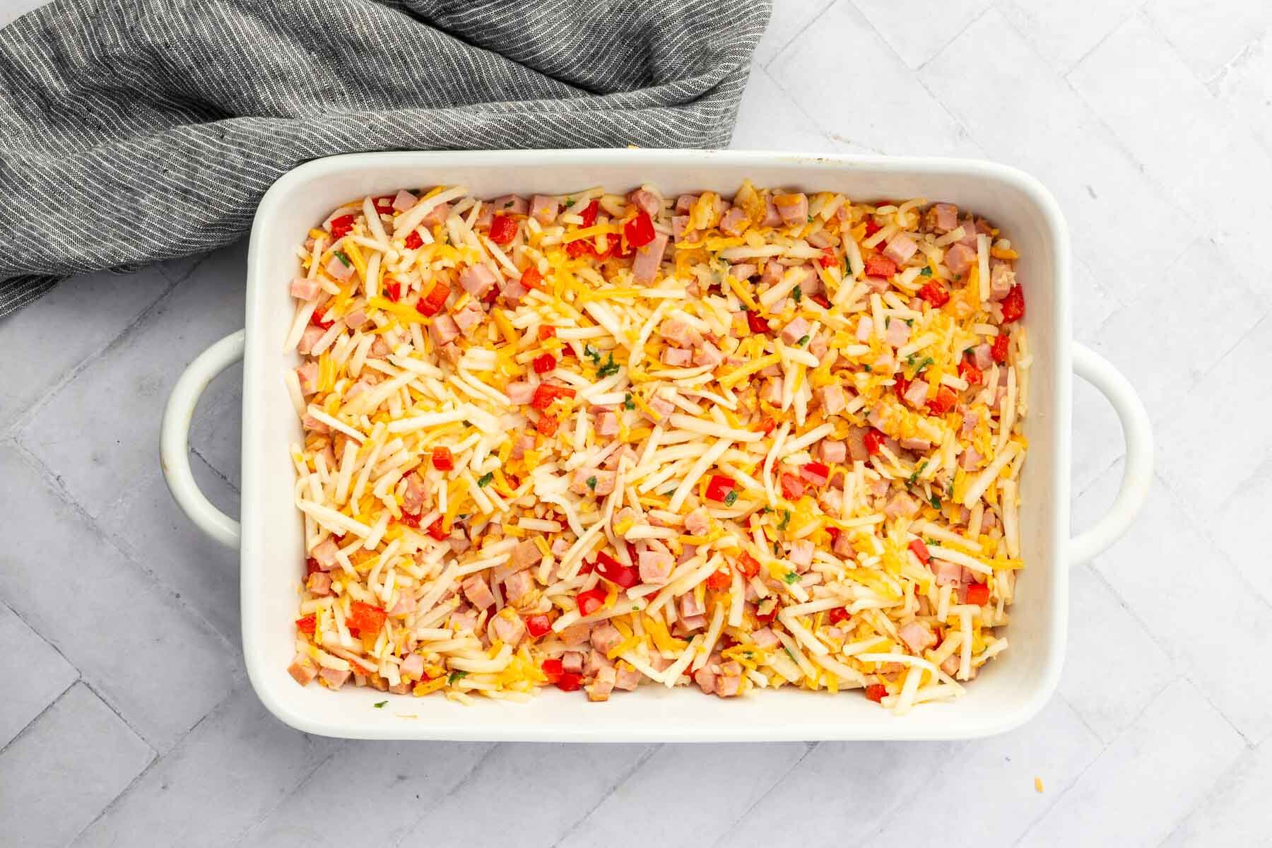 A white baking dish with hash browns, cheese, cubed ham, diced bell pepper before baking in the oven.