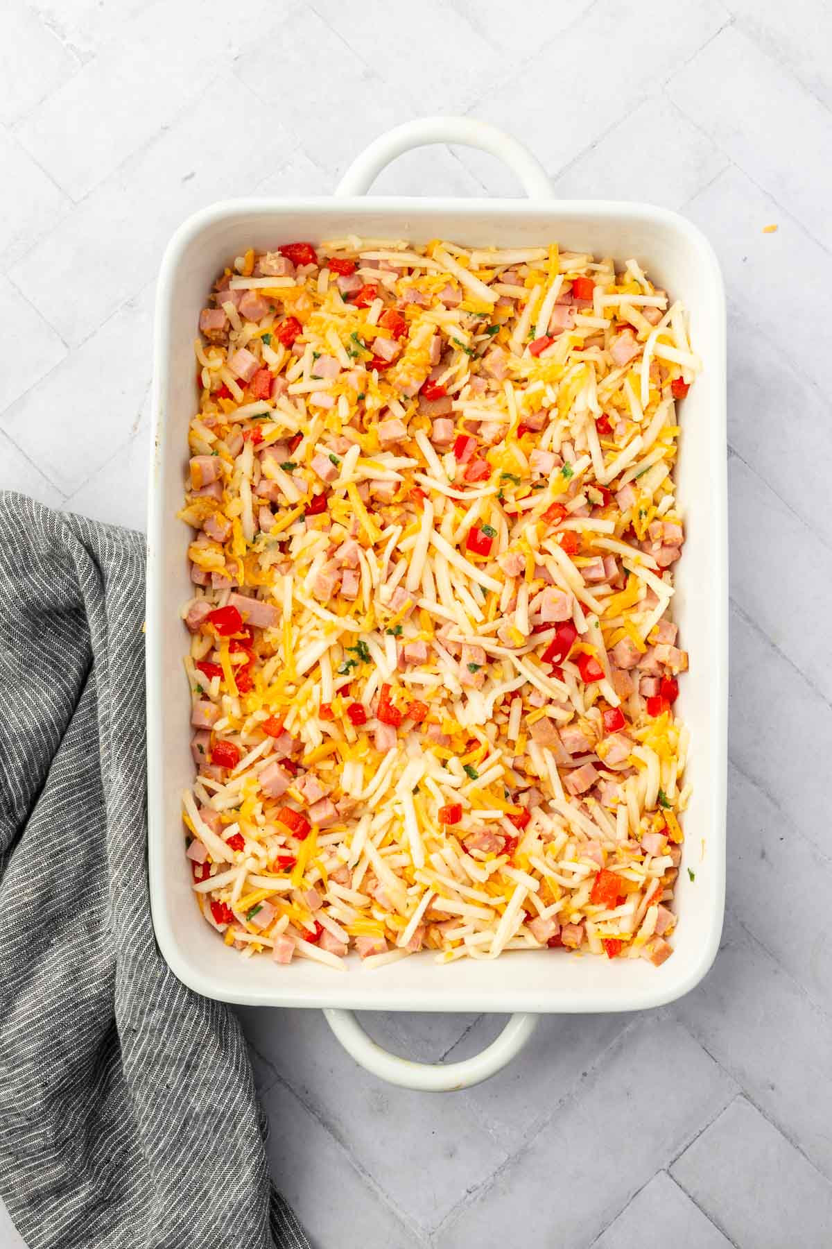 A white baking dish with hash browns, cheese, cubed ham, diced bell pepper before baking in the oven.
