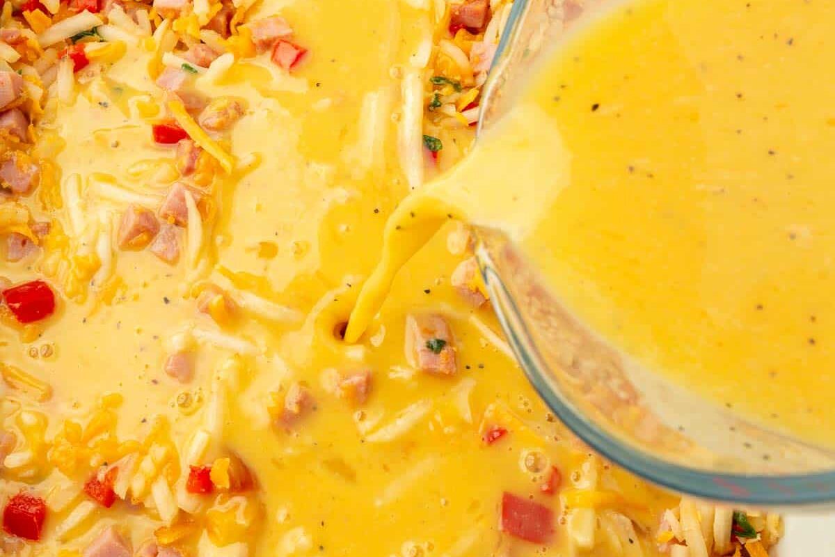 A measuring cup pouring an egg mixture over hash browns, red bell pepper, cubed ham and cheese in a baking dish.
