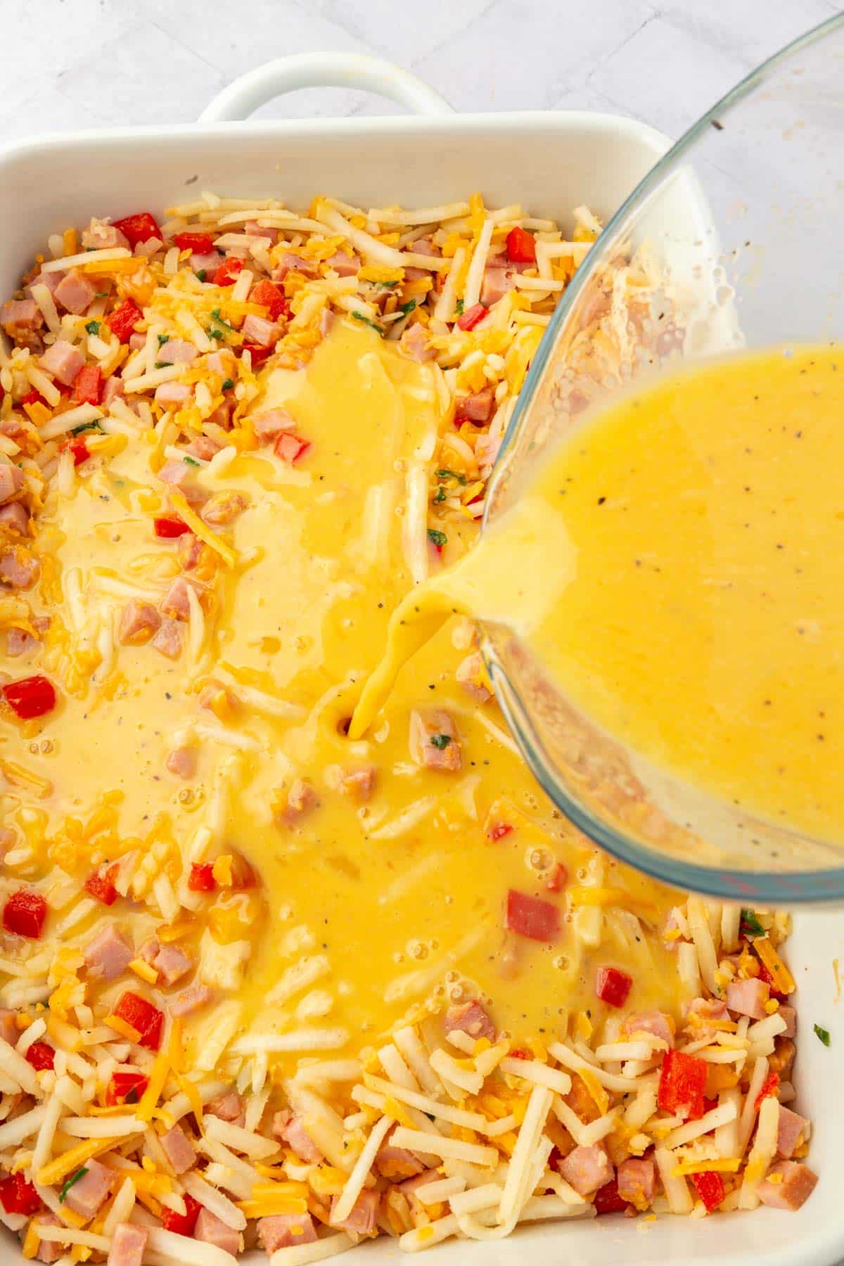 A measuring cup pouring an egg mixture over hash browns, red bell pepper, cubed ham and cheese in a baking dish.