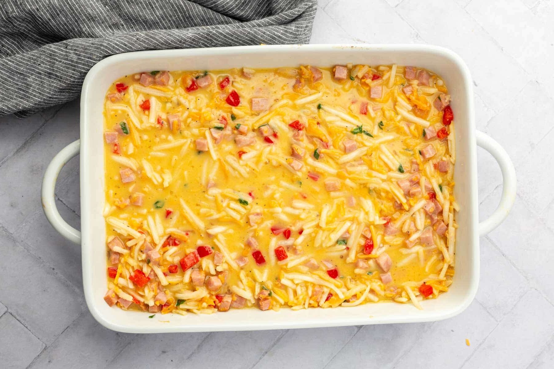 A 9x13-inch white baking dish with hash browns, eggs, cubed ham, cheese, red pepper, and green onions before baking in the oven.