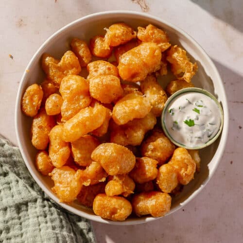 An overhead view of a bowl of gluten-free cheese curds that have been deep fried with a small ramekin of ranch dressing and another ramekin of marinara sauce on the side.