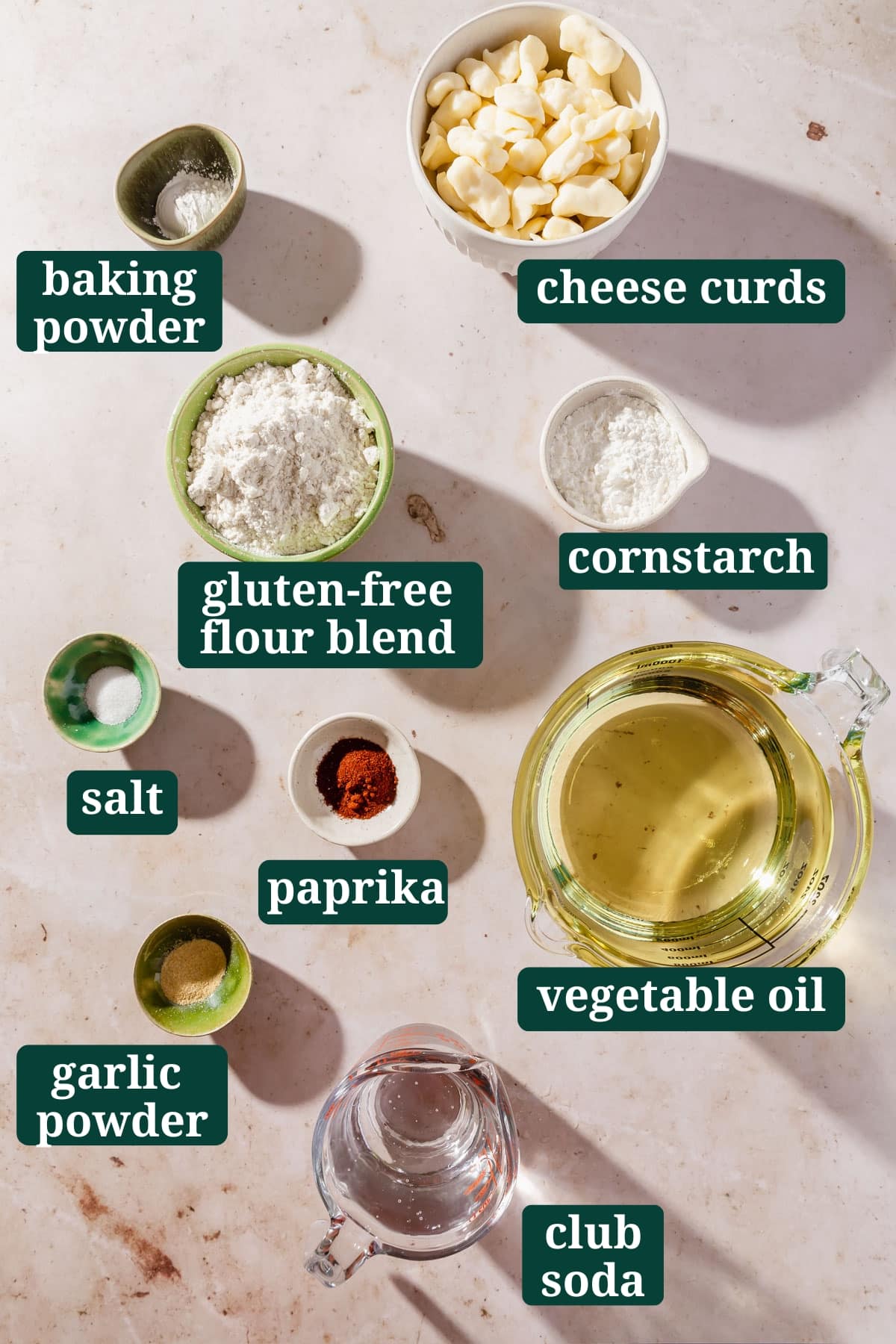 Ingredients in small bowls on a table to make gluten-free cheese curds, including white cheddar cheese curds, baking powder, gluten-free flour, cornstarch, salt, paprika, vegetable oil, garlic powder, and club soda, with text overlays over each ingredient.