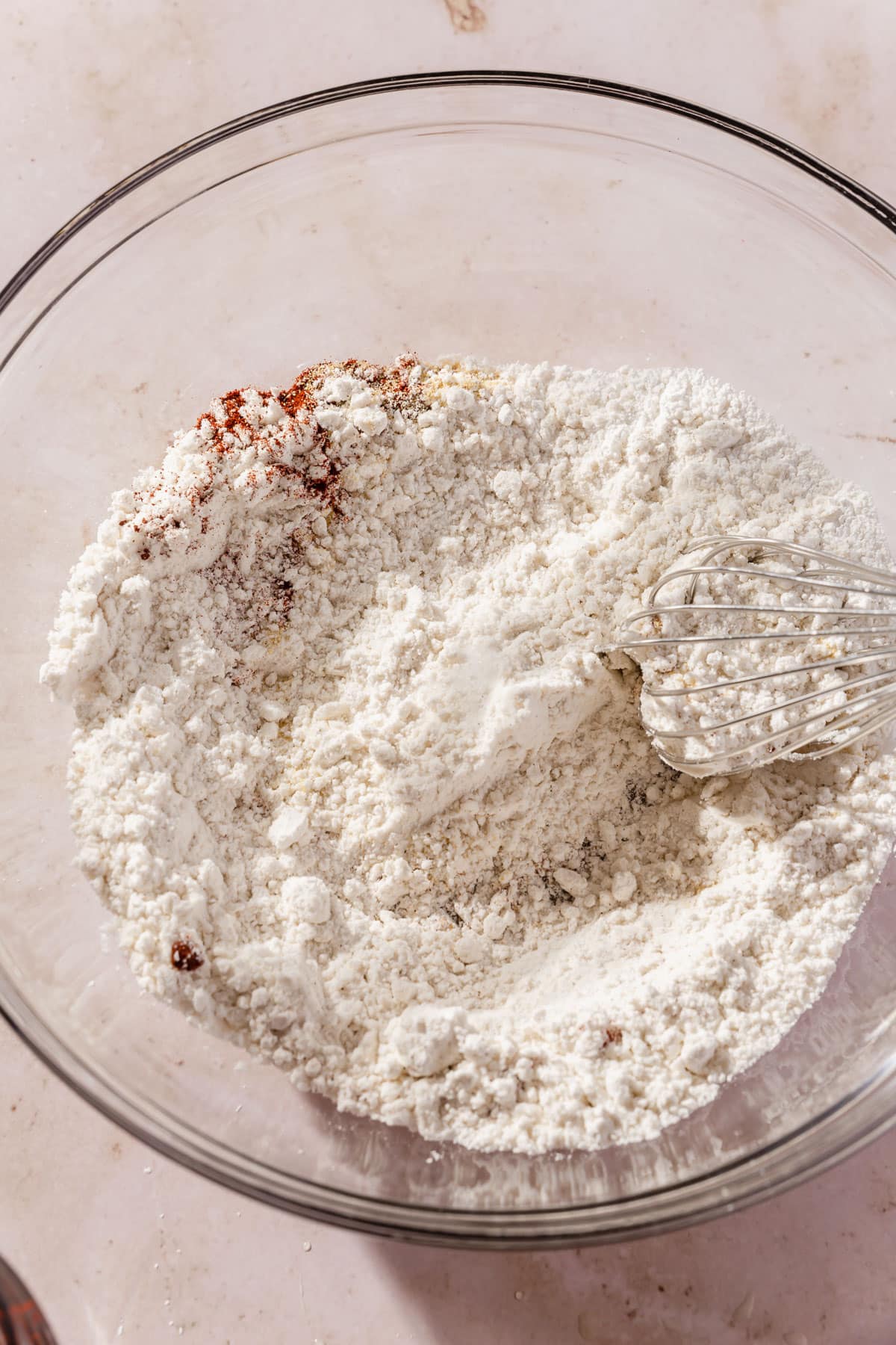A glass mixing bowl of gluten-free flour, paprika, salt, garlic powder, and baking powder being mixed together with a whisk.