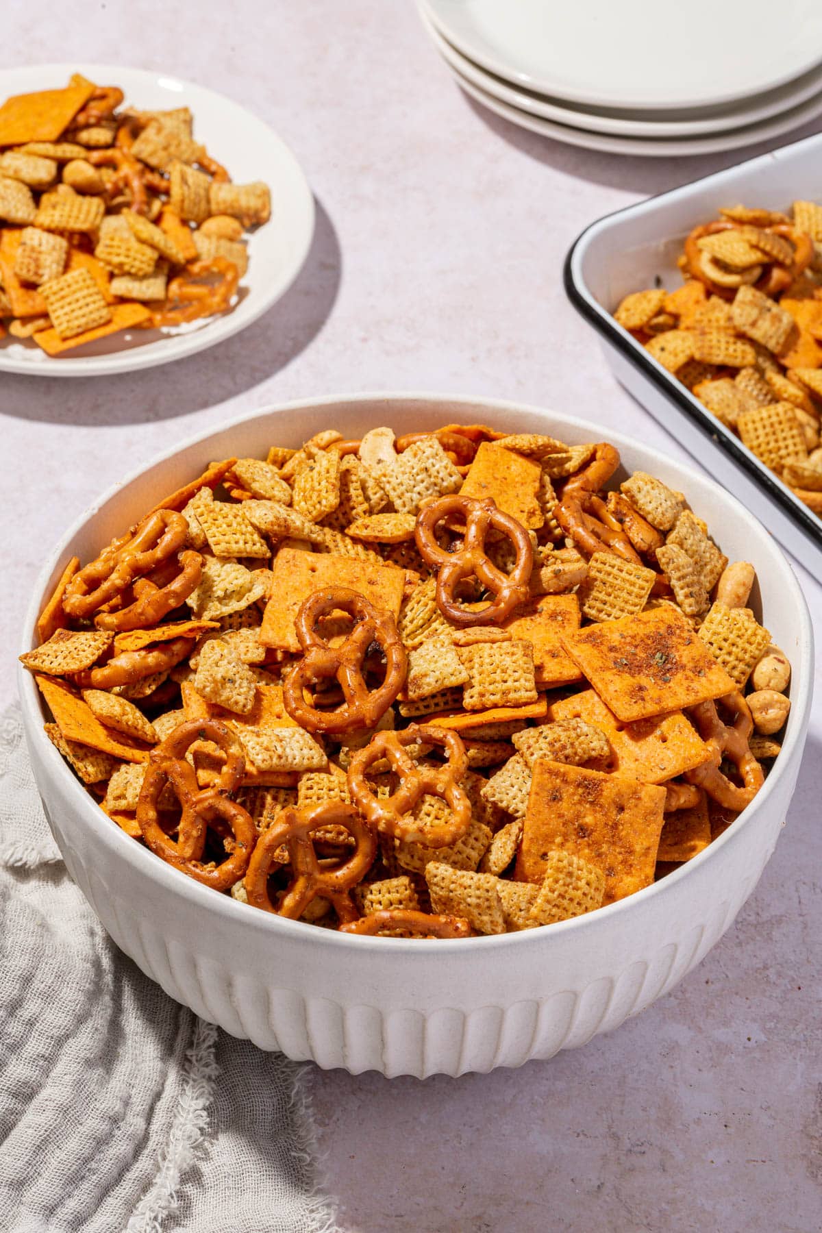 A serving bowl of gluten-free chex mix with a tray of more chex mix and a stack of appetizer plates in the background.