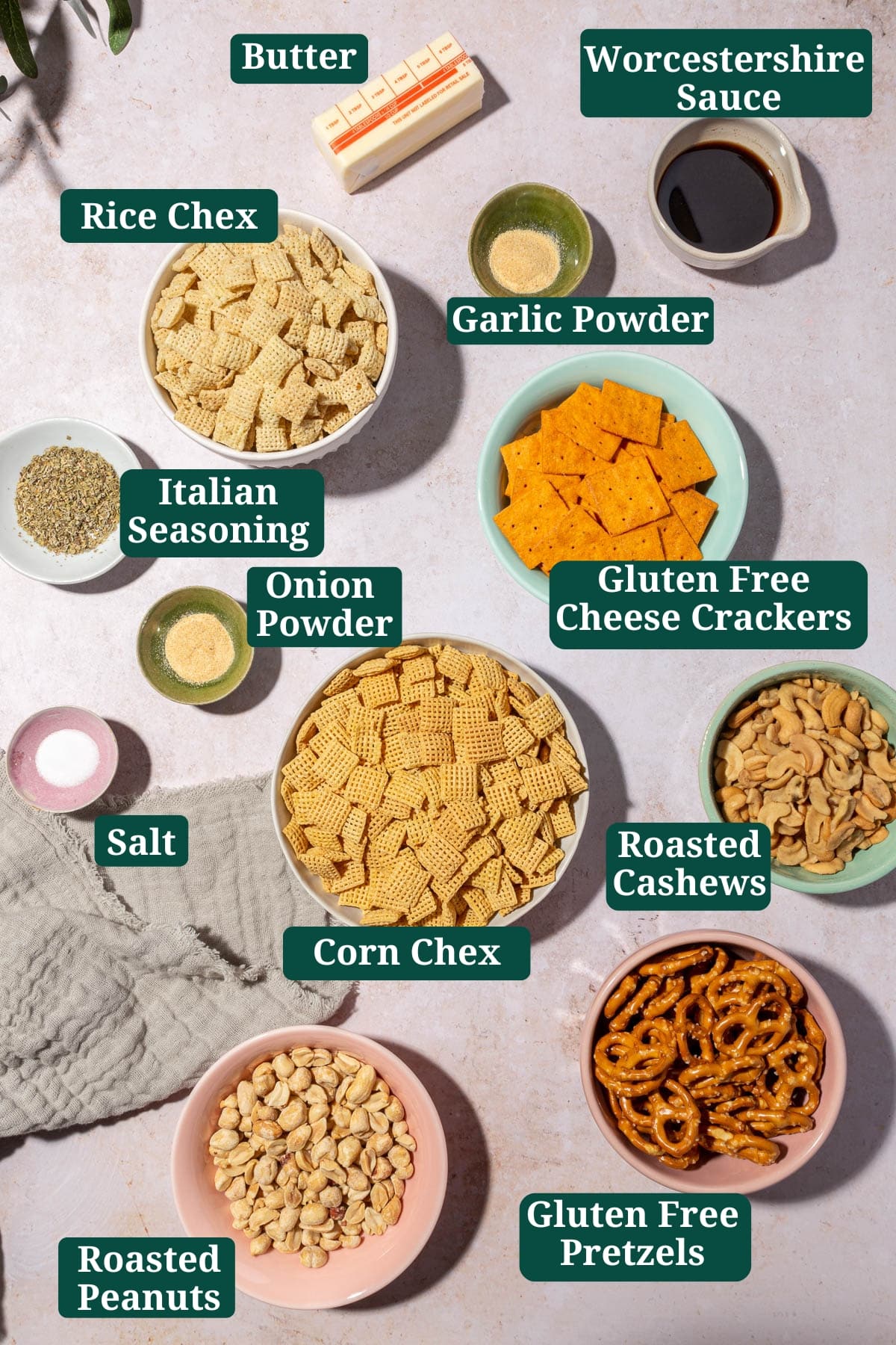 An overhead view of ingredients in small bowls to make gluten-free chex mix at home including corn chex, rice chex, gluten-free pretzels, gluten-free cheese crackers, cashews, peanuts, butter, garlic powder, onion powder, salt, and Italian seasoning with text overlays over each ingredient.