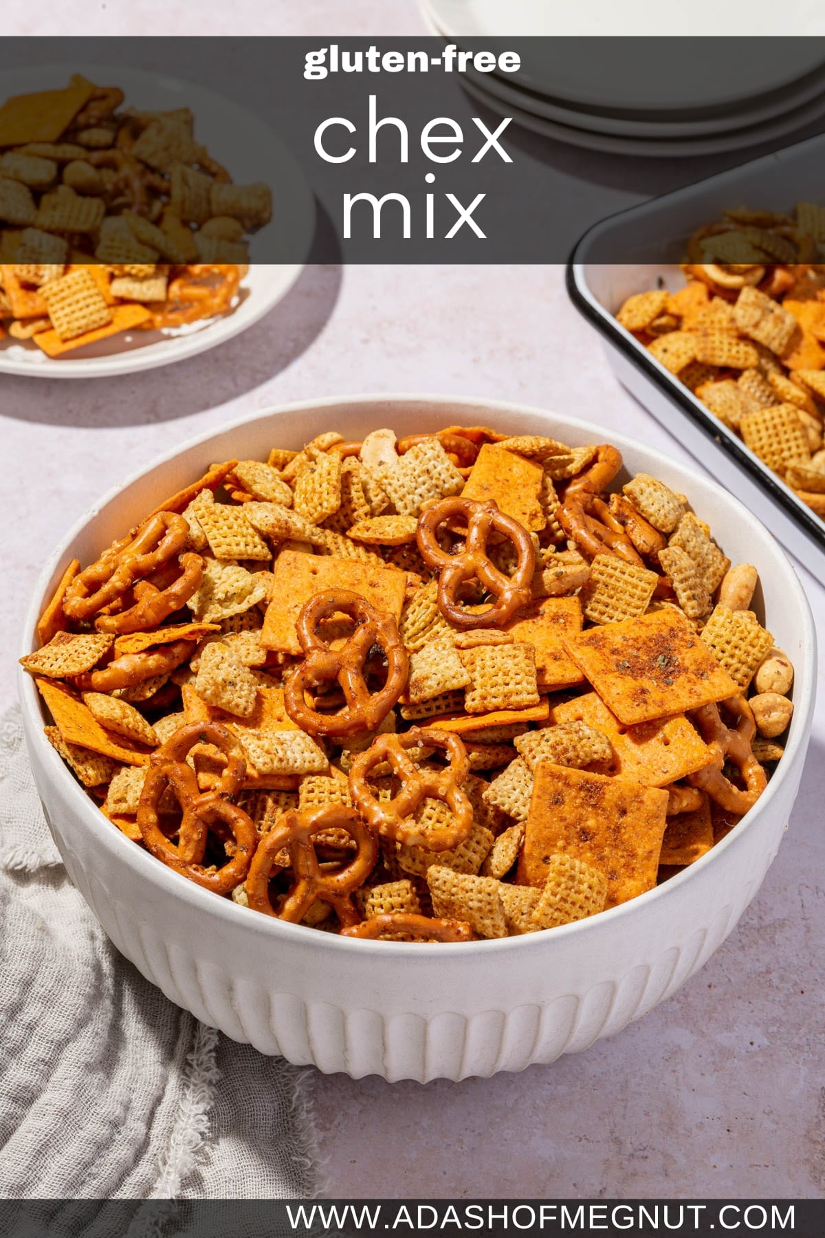 A serving bowl of gluten-free chex mix with a tray of more chex mix and a stack of appetizer plates in the background.