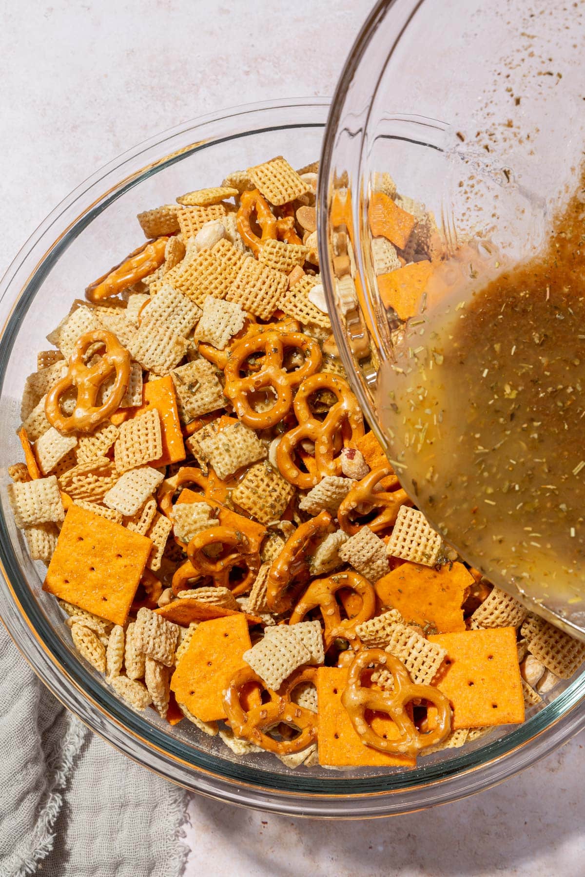 A bowl of melted butter and spice mixture being drizzled over gluten-free chex mix in a glass bowl.