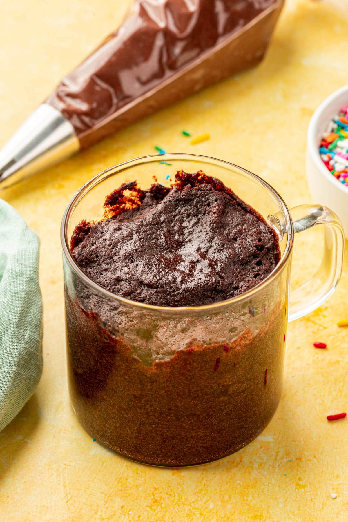 A glass mug of gluten-free chocolate mug cake with a piping bag of chocolate buttercream in the background.