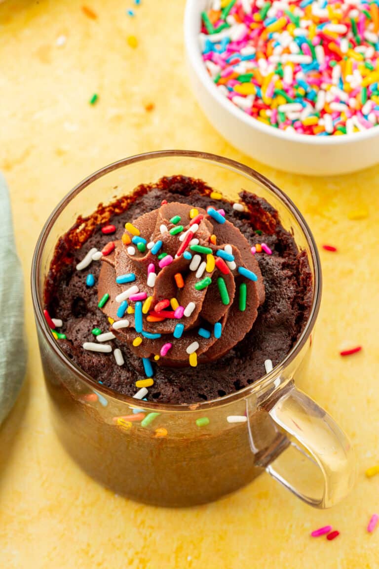 A closeup of a gluten-free mug cake in a glass mug topped with a swirl of chocolate buttercream and colorful sprinkles.