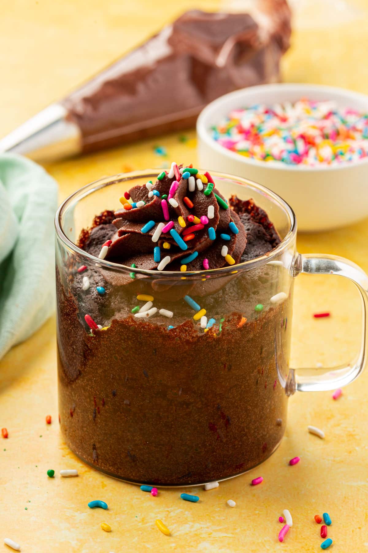 A chocolate mug cake in a glass mug topped with chocolate frosting and rainbow sprinkles.