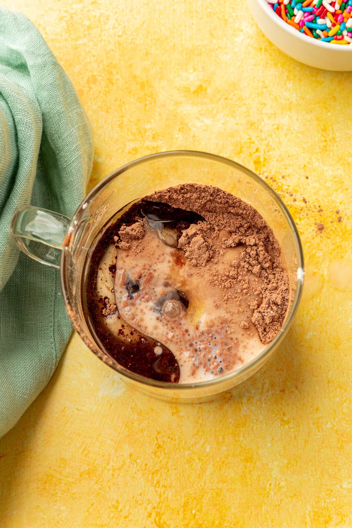 Cocoa powder, almond milk, oil, and vanilla in a glass mug before mixing together to make a gluten-free mug cake.