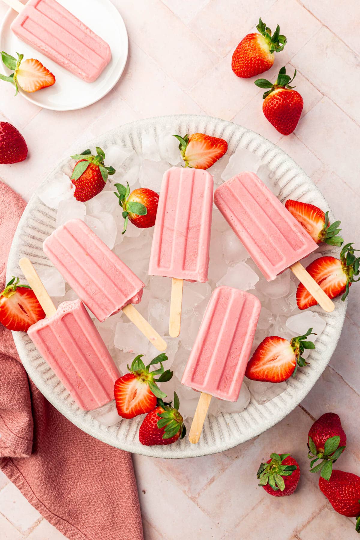 A large bowl filled with ice cubes and topped with strawberry yogurt popsicles with fresh strawberry halves surrounding the popsicles.