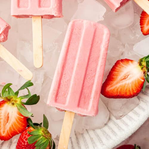 A closeup of a strawberry popsicle in a large bowl of of ice with fresh strawberry halves surrounding the popsicles.