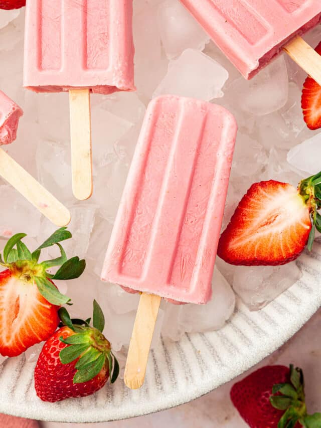 A closeup of a strawberry popsicle in a large bowl of of ice with fresh strawberry halves surrounding the popsicles.