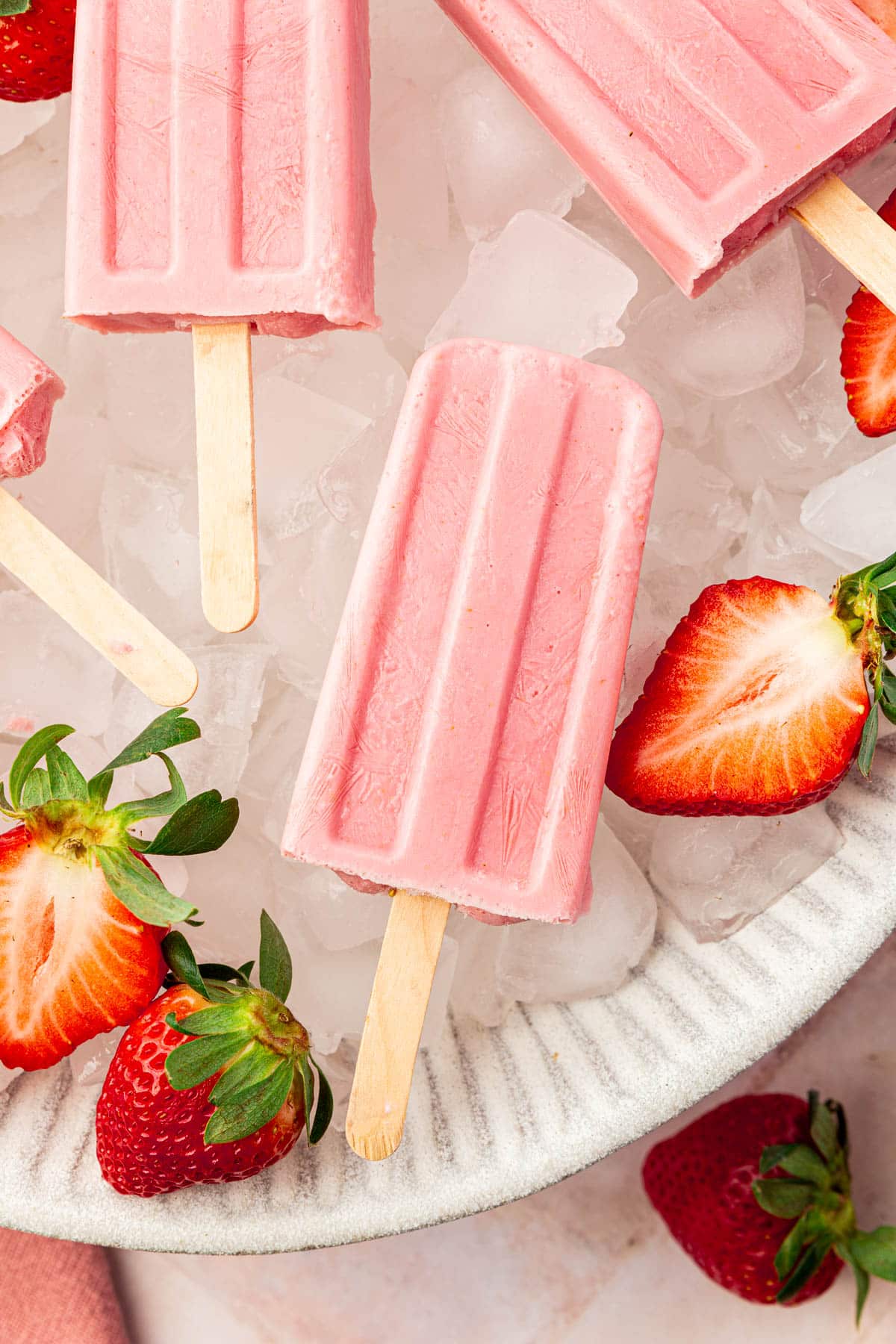 A closeup of a strawberry popsicle in a large bowl of of ice with fresh strawberry halves surrounding the popsicles.