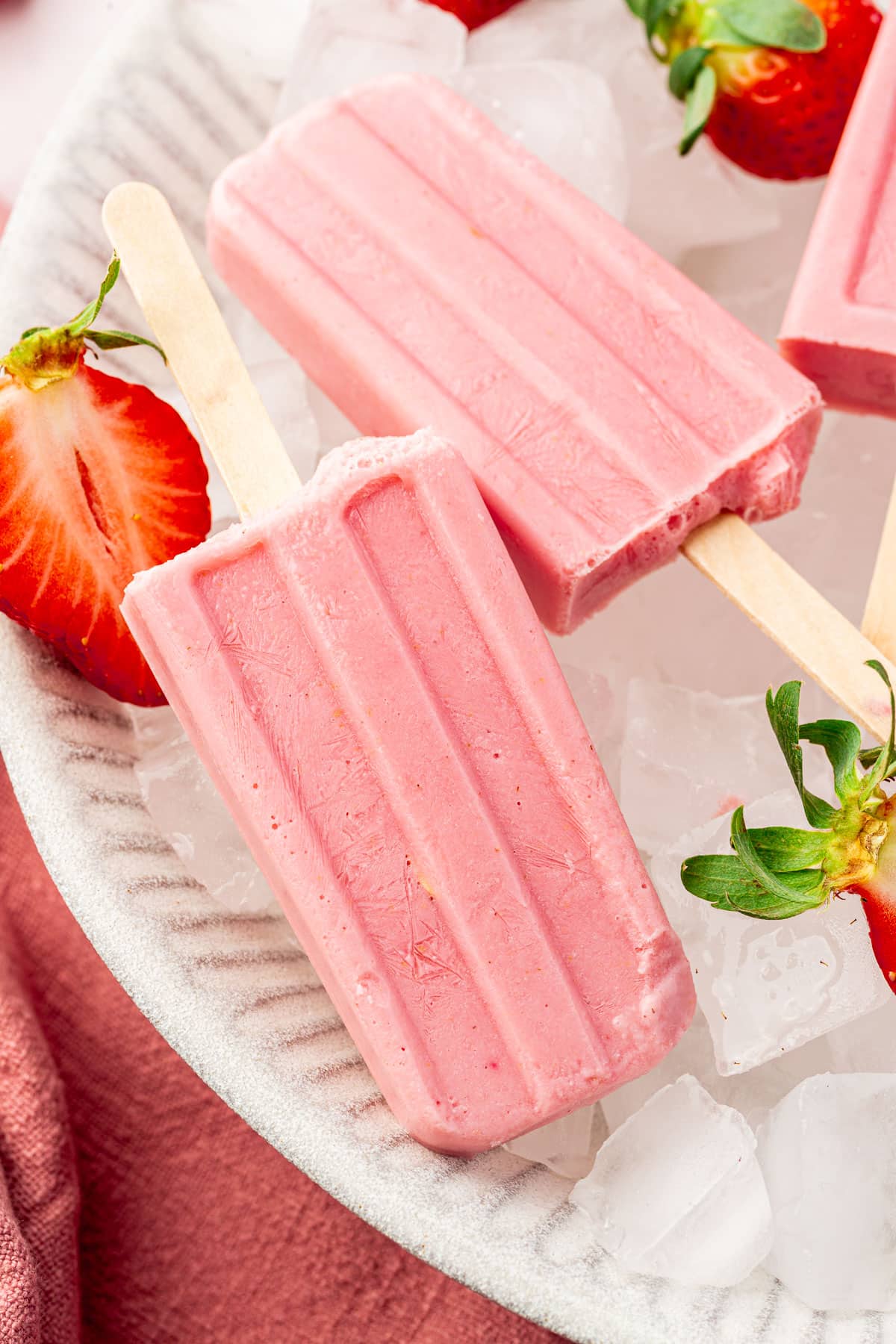 Two strawberry popsicles in a large bowl of ice with fresh strawberry halves surrounding the popsicles.