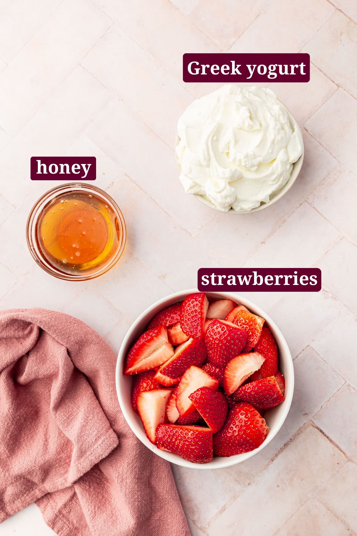 A bowl of strawberry quarters, a small bowl of honey, and a medium bowl of Greek yogurt on a pink table with text overlays over each ingredient.