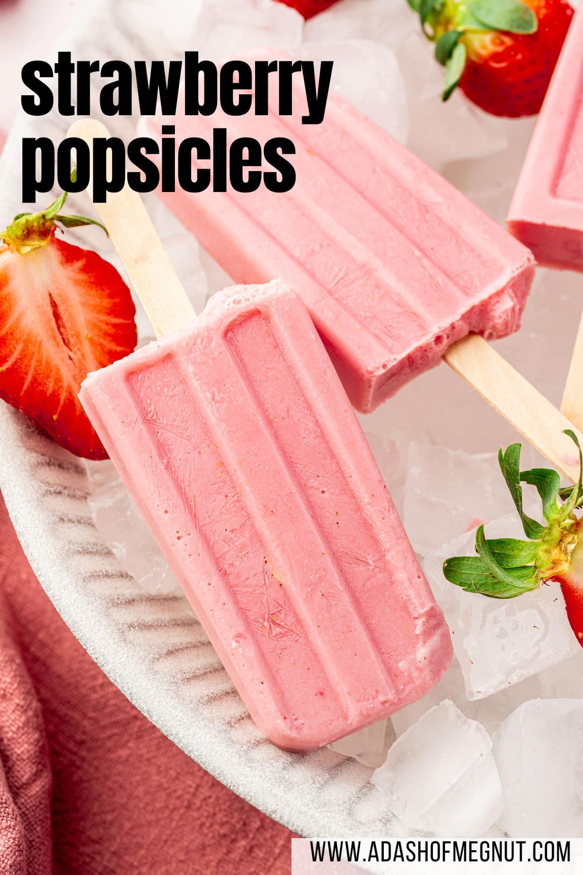 A close up of a strawberry popsicle laying in a large bowl of ice cubes with some fresh strawberry halves surrounding the popsicles.