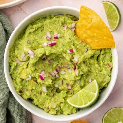 An overhead view of a bowl of guacamole topped with diced red onion, a tortilla chip and a lime wedge with additional chips and limes on the surface.