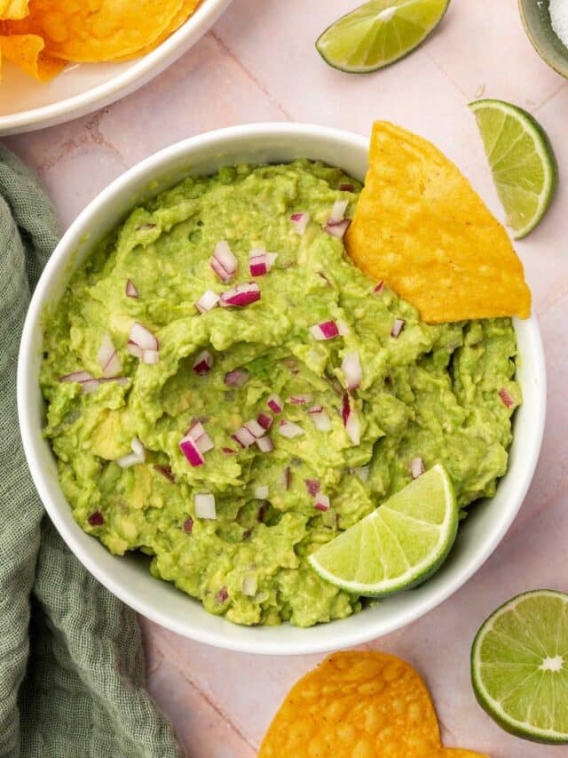 An overhead view of a bowl of guacamole topped with diced red onion, a tortilla chip and a lime wedge with additional chips and limes on the surface.