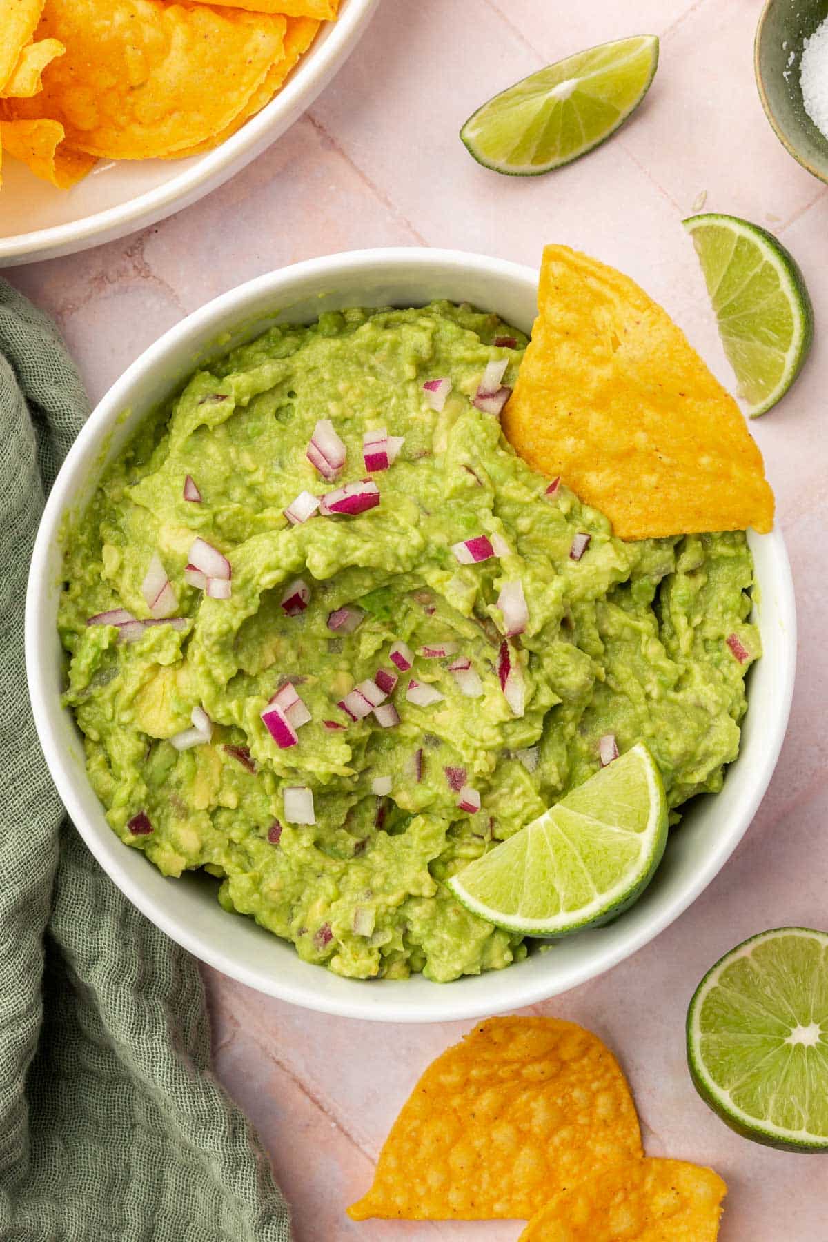 An overhead view of a bowl of guacamole topped with diced red onion, a tortilla chip and a lime wedge with additional chips and limes on the surface.