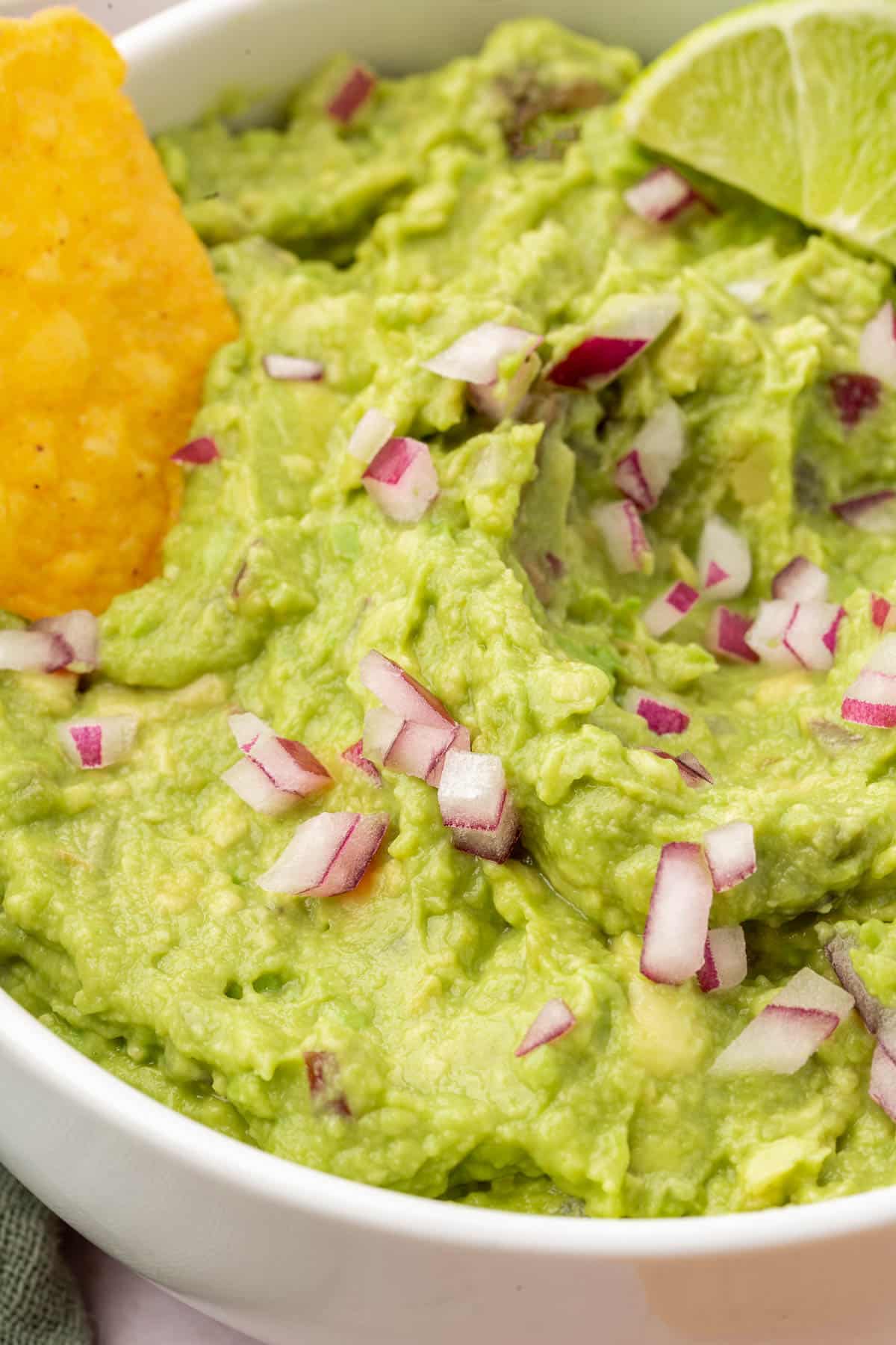 A closeup of a bowl of guacamole topped with diced red onion, a lime wedge and a single tortilla chip.