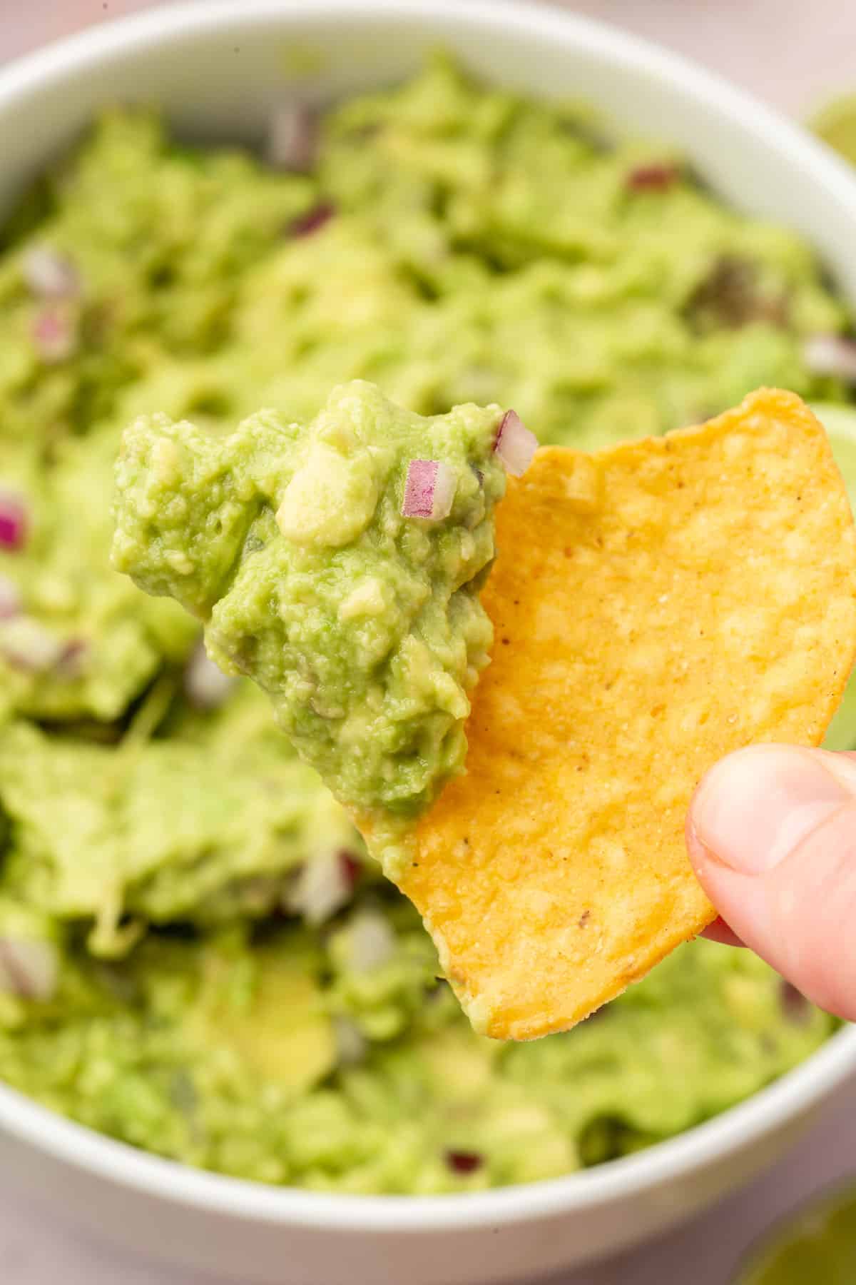 A hand holding a tortilla chip dipped in guacamole over a bowl of more guacamole.