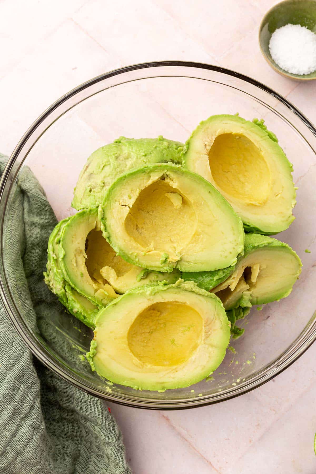 A glass mixing bowl with six avocado halves in it with a bowl of kosher salt to the side.