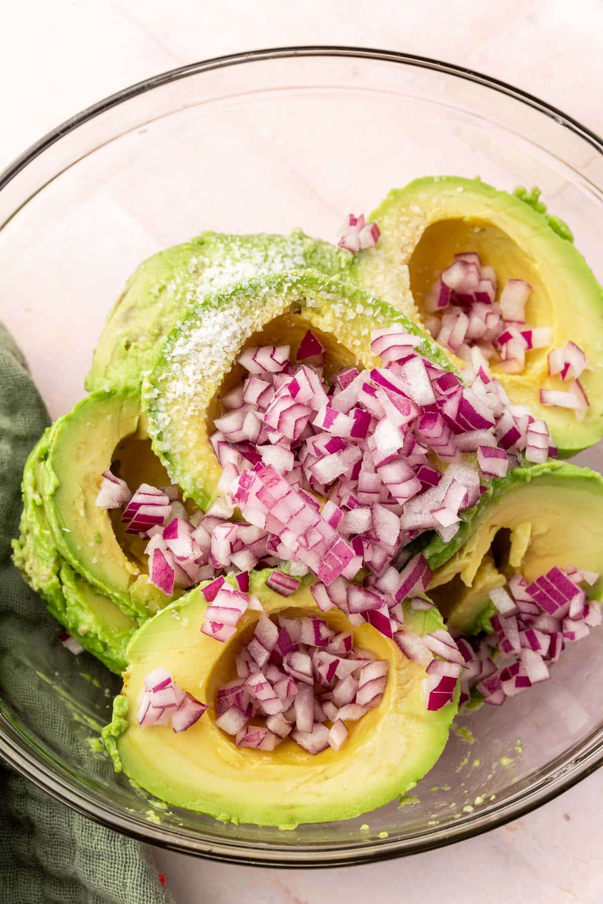 A glass mixing bowl with avocado halves, salt, lime juice and finely diced red onion in it.