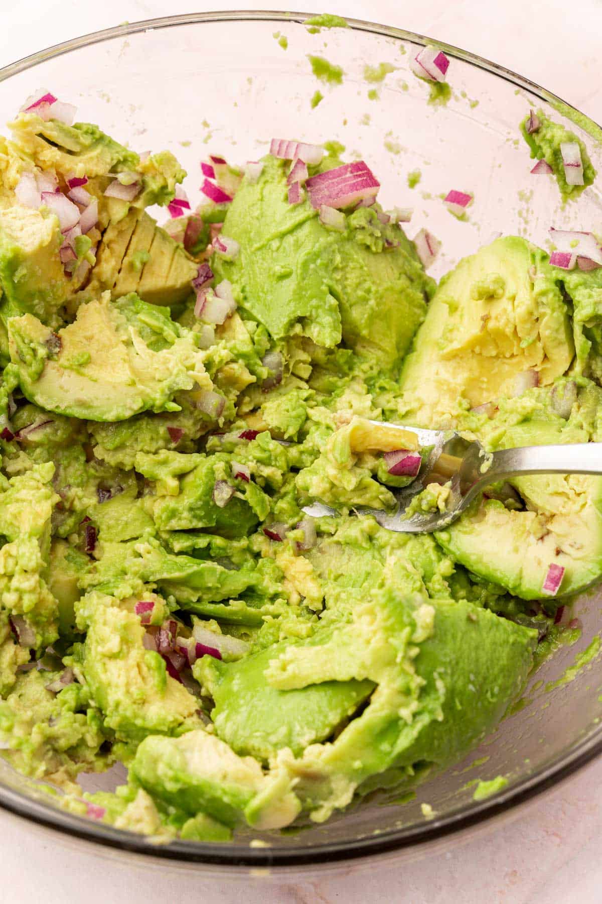 A closeup of avocados and red onion being mashed with a fork in a glass mixing bowl.
