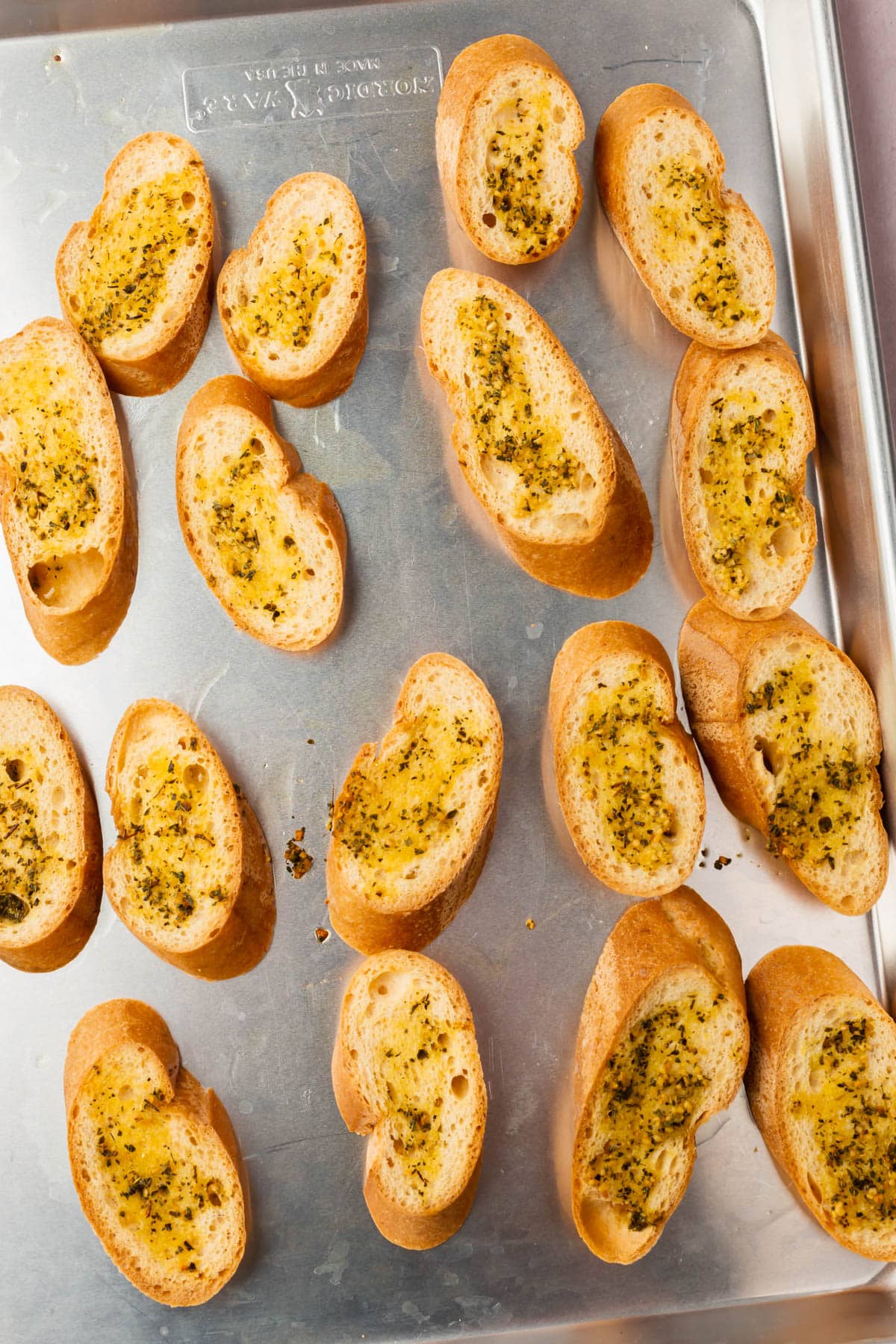 A sheet pan with slices of gluten-free bread cut on the bias and topped with olive oil, minced garlic and dried herbs.