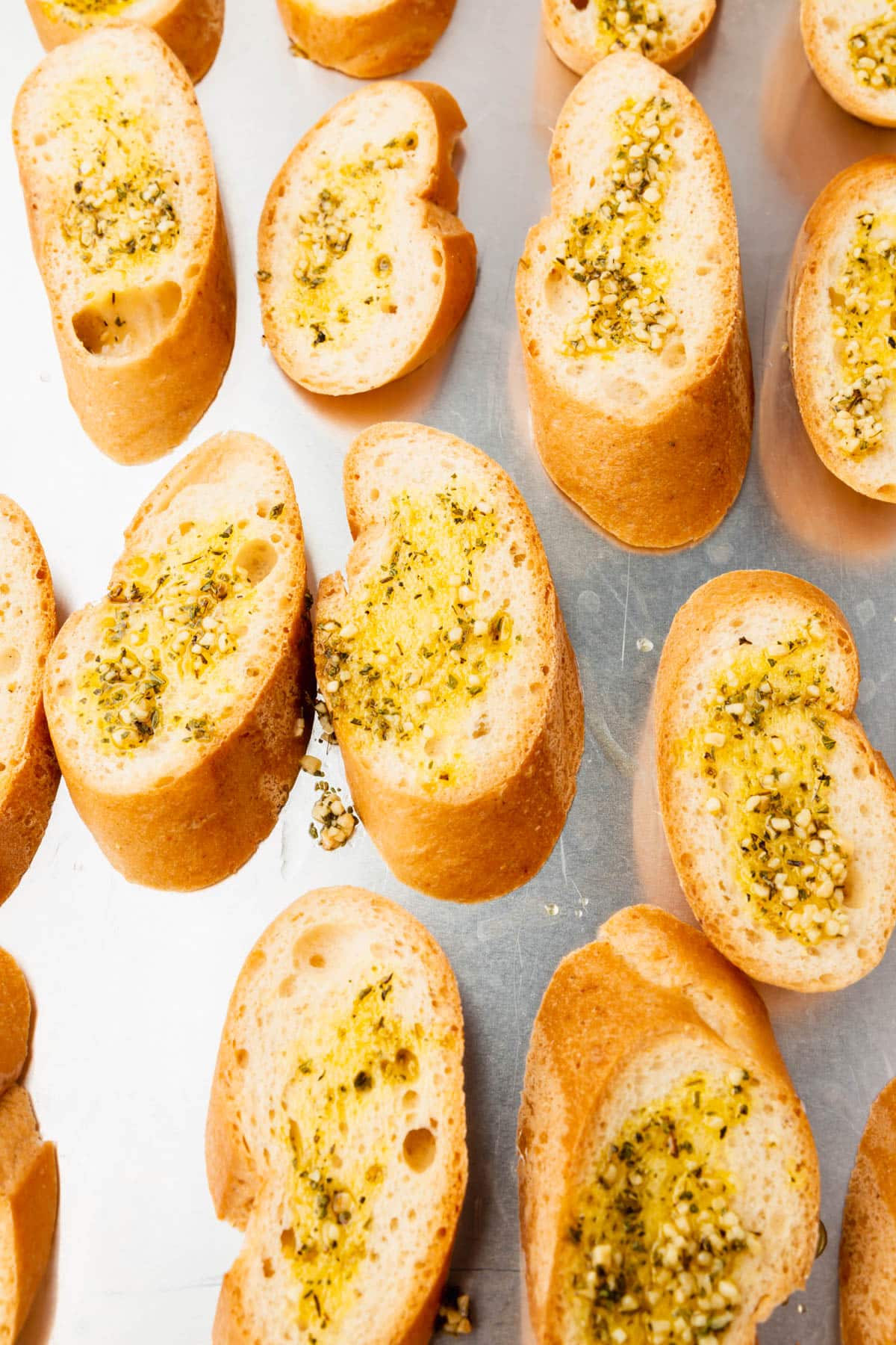 A close up of gluten free bread on a baking sheet topped with olive oil, minced garlic and dried herbs before toasting in the oven.